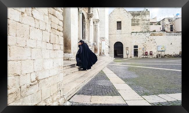 Bari, Italy - March 12, 2019: A Catholic priest walks towards the cathedral dressed in a black cape and hat a day of wind and rain. Framed Print by Joaquin Corbalan