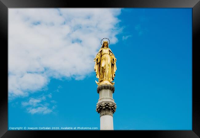 Golden religious statue, illuminated by the sun, of the Virgin Mary on top of a pedestal, with a background of blue sky and clouds. Framed Print by Joaquin Corbalan