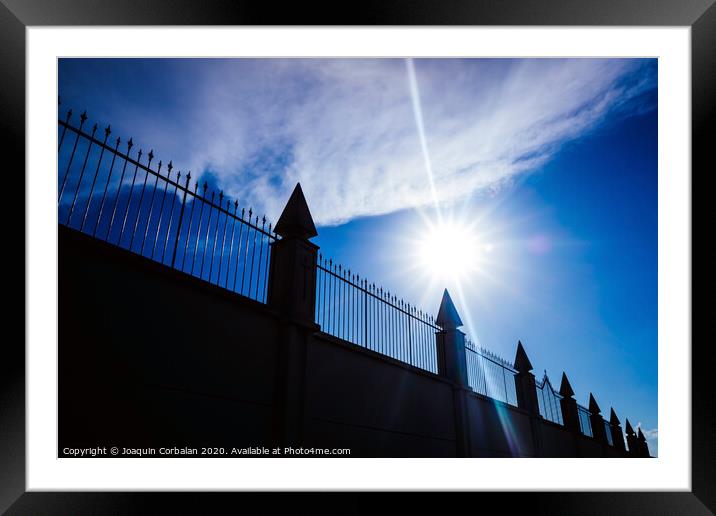 Silhouette against the sun of a high wall and metal fence with an intense blue sky in the background. Framed Mounted Print by Joaquin Corbalan