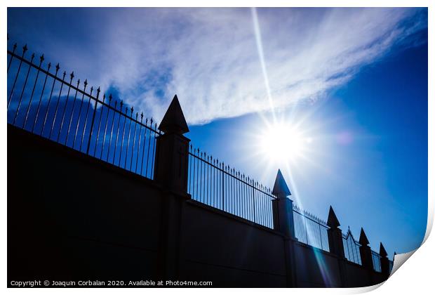 Silhouette against the sun of a high wall and metal fence with an intense blue sky in the background. Print by Joaquin Corbalan