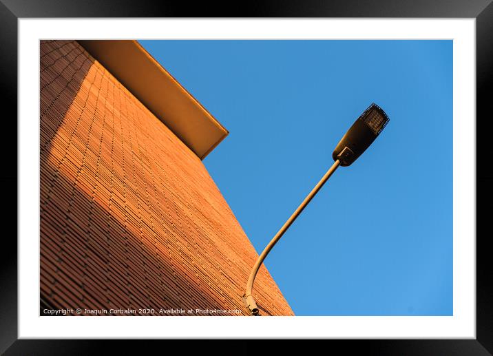 A deep red brick wall supports a lamppost, contrasting and bright colors. Framed Mounted Print by Joaquin Corbalan