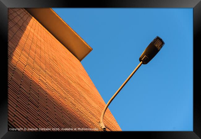 A deep red brick wall supports a lamppost, contrasting and bright colors. Framed Print by Joaquin Corbalan