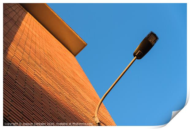 A deep red brick wall supports a lamppost, contrasting and bright colors. Print by Joaquin Corbalan