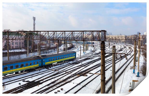Passenger railway wagons ride along the railway tracks in the winter season against the backdrop of the cityscape Print by Sergii Petruk