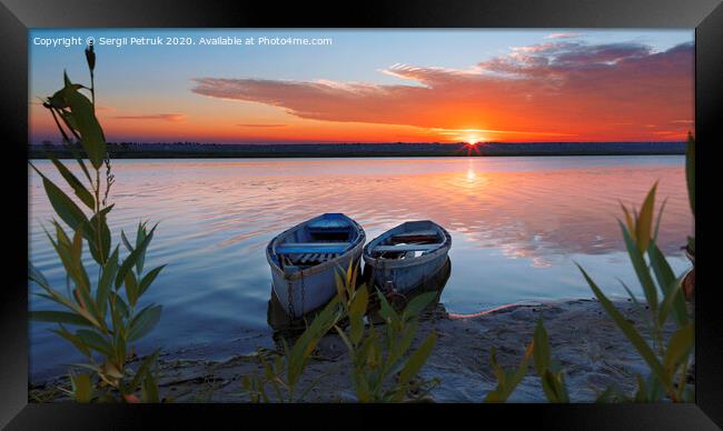 Two boats stand at the shore of a calm river against the background of a bright rising sun Framed Print by Sergii Petruk