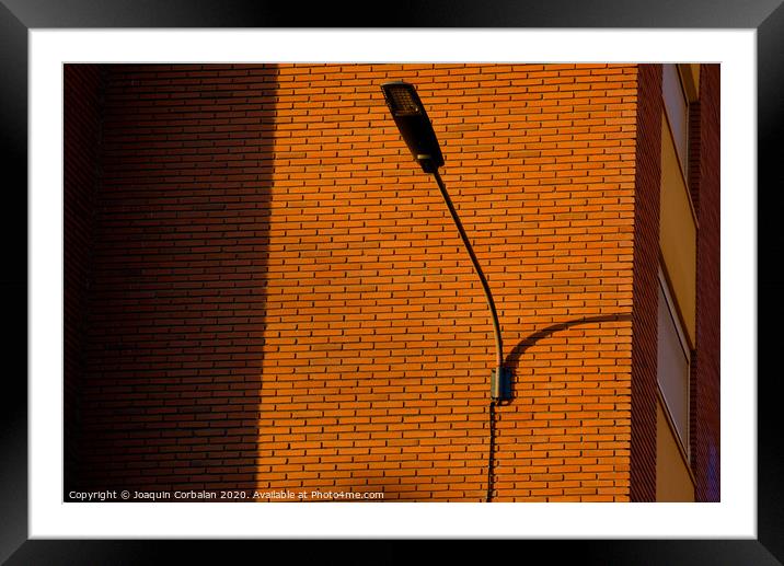 A deep red brick wall supports a lamppost, contrasting and bright colors. Framed Mounted Print by Joaquin Corbalan