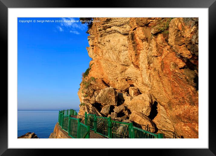 The road along the sea coast, surrounded by a green metal fence, is illuminated by the rays of the morning golden sun near a rocky cliff. Framed Mounted Print by Sergii Petruk