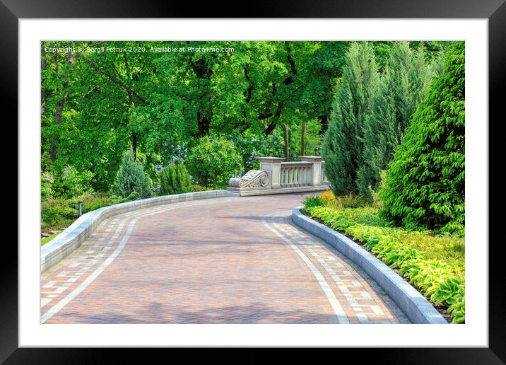 Walkway paved with tiles in a beautiful park, framed by different bushes and flowers, in soft rays of morning light. Framed Mounted Print by Sergii Petruk
