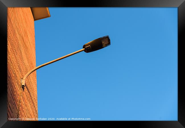 Lamppost on the red wall of a building, by day, with a blue sky background. Framed Print by Joaquin Corbalan
