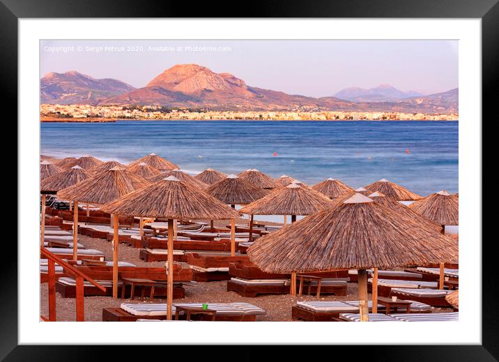 The straw tops of beach umbrellas and wooden deck chairs with mattresses on the deserted beach promenade in the rays of the evening setting sun. Framed Mounted Print by Sergii Petruk