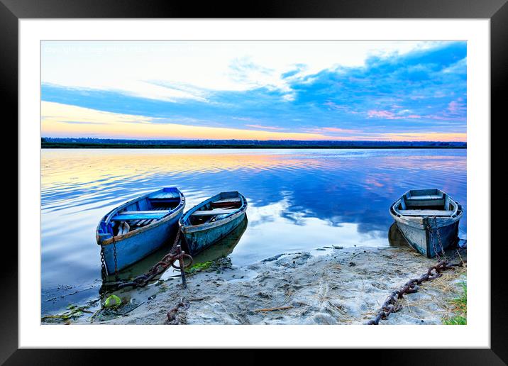 Old blue-green boats moored to the shore by old iron chains and stand on the shore of a calm river against the background of the morning horizon. Framed Mounted Print by Sergii Petruk