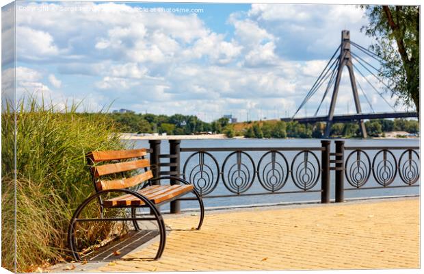 A wooden bench on the embankment of the Dnipro River against the background of yellow paving slabs and the northern bridge over the river in blur. Canvas Print by Sergii Petruk