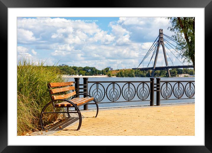 A wooden bench on the embankment of the Dnipro River against the background of yellow paving slabs and the northern bridge over the river in blur. Framed Mounted Print by Sergii Petruk