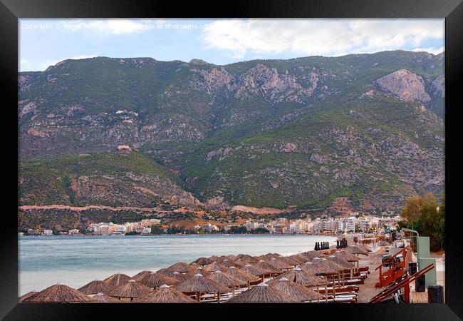 View of thatched peaks of beach umbrellas and wooden sun loungers with mattresses on the deserted waterfront of Loutraki in Greece against the backdrop of mountains in the early morning. Framed Print by Sergii Petruk