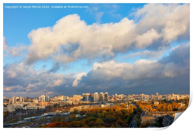 The colorful cityscape of autumn Kyiv, the sun's rays from behind gray clouds catch and brightly illuminate the orange foliage of the city park. Print by Sergii Petruk