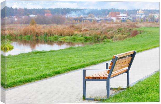 A wooden bench stands on a walking path along a beautiful green lawn on the river embankment with a blurred background of a rural landscape. Canvas Print by Sergii Petruk