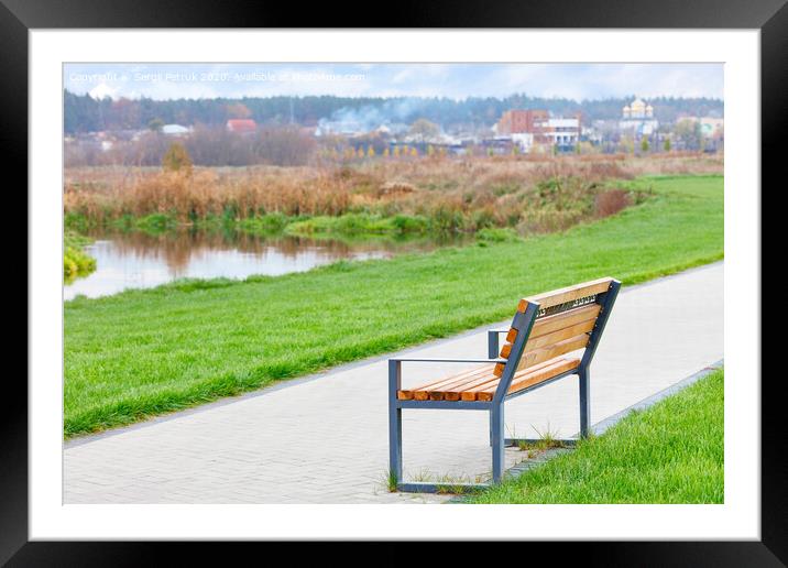 A wooden bench stands on a walking path along a beautiful green lawn on the river embankment with a blurred background of a rural landscape. Framed Mounted Print by Sergii Petruk