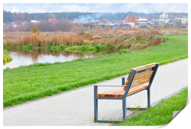 A wooden bench stands on a walking path along a beautiful green lawn on the river embankment with a blurred background of a rural landscape. Print by Sergii Petruk