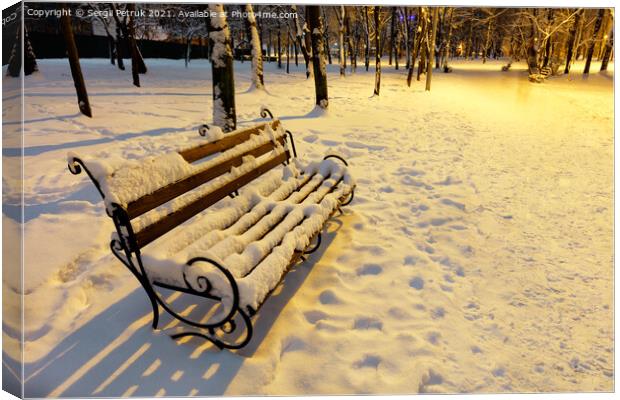 A wooden bench in a winter city evening park is covered with snow and illuminated by the warm light of a street lamp. Canvas Print by Sergii Petruk