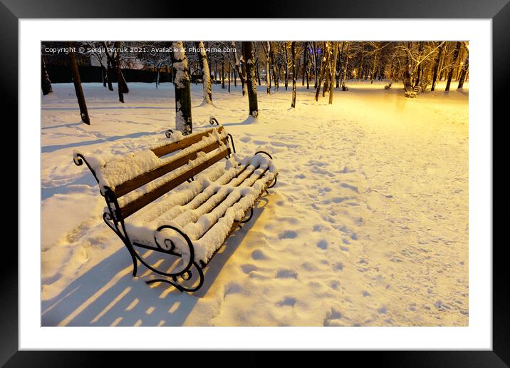 A wooden bench in a winter city evening park is covered with snow and illuminated by the warm light of a street lamp. Framed Mounted Print by Sergii Petruk