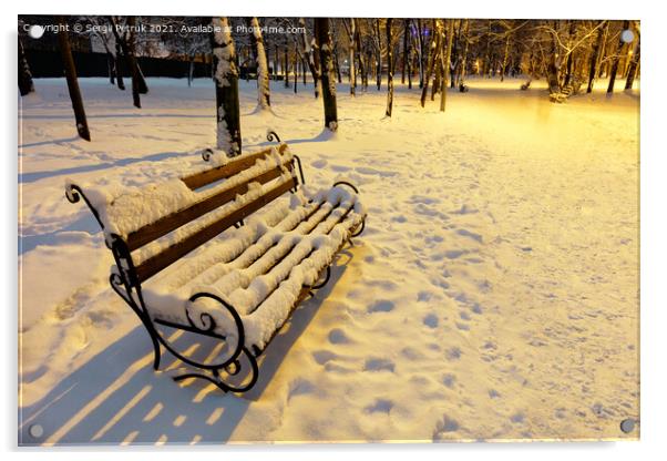A wooden bench in a winter city evening park is covered with snow and illuminated by the warm light of a street lamp. Acrylic by Sergii Petruk