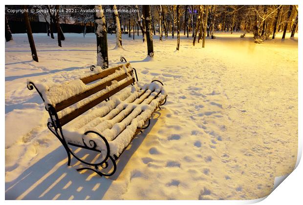 A wooden bench in a winter city evening park is covered with snow and illuminated by the warm light of a street lamp. Print by Sergii Petruk