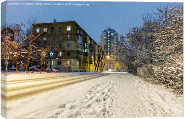 Snow-covered road along a city street with trees in the snow and city evening lighting against the backdrop of blue twilight. Canvas Print by Sergii Petruk