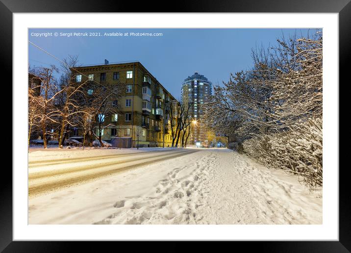 Snow-covered road along a city street with trees in the snow and city evening lighting against the backdrop of blue twilight. Framed Mounted Print by Sergii Petruk