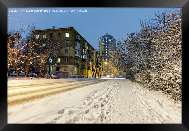 Snow-covered road along a city street with trees in the snow and city evening lighting against the backdrop of blue twilight. Framed Print by Sergii Petruk