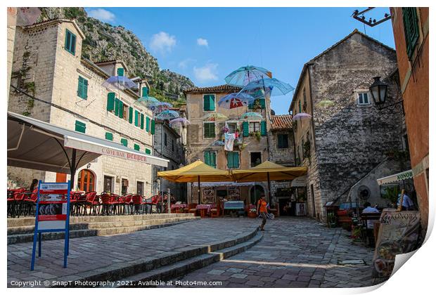 Cafe's and flying umbrellas on a square in the old town of Kotor, Montenegro Print by SnapT Photography