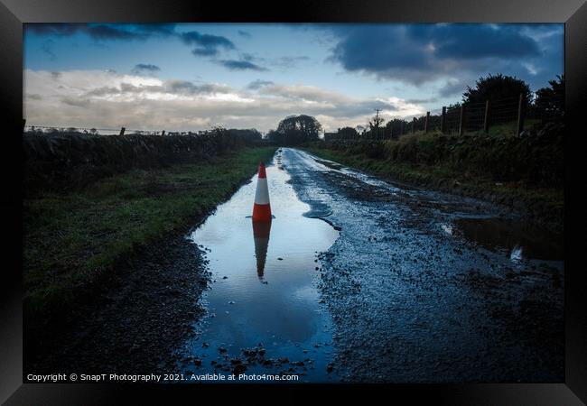 A red traffic cone reflecting a pot hole containing a pool of water on a road Framed Print by SnapT Photography