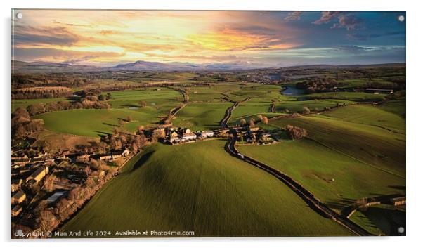 Aerial view of a scenic countryside at sunset with lush green fields, a small village, and a winding road leading towards distant hills. Acrylic by Man And Life