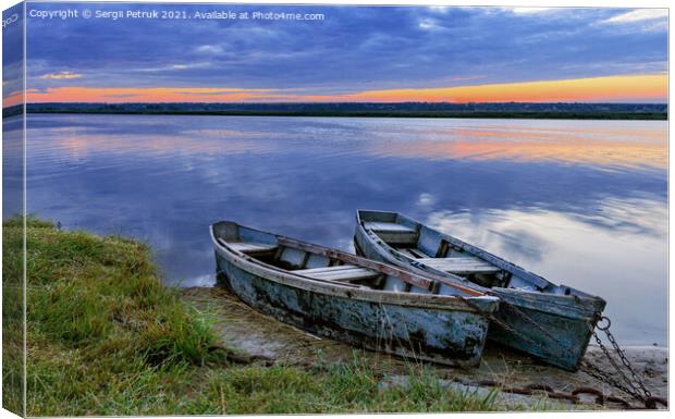 Two old dilapidated boats are moored with a metal chain to the bank of a calm river in the early morning against the backdrop of a cloudy sky and the rising sun. Canvas Print by Sergii Petruk