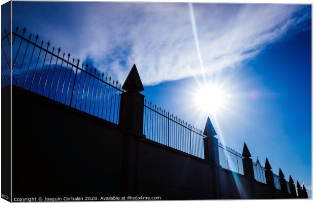 Silhouette against the sun of a high wall and metal fence with an intense blue sky in the background. Canvas Print by Joaquin Corbalan