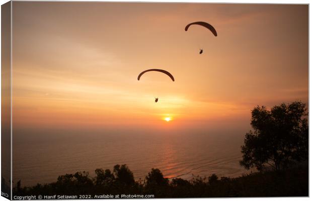 Two paraglider over treetops and ocean at sunset Canvas Print by Hanif Setiawan