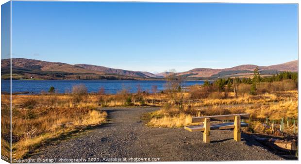 A wooden bench overlooking Clatteringshaws Loch on a sunny winters day Canvas Print by SnapT Photography