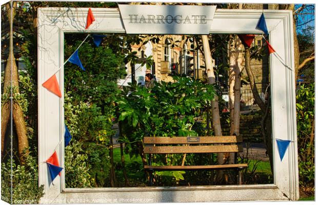 Quaint garden scene framed by a white wooden structure with 'Harrogate' sign, featuring a bench and lush greenery, adorned with colorful pennants. Canvas Print by Man And Life