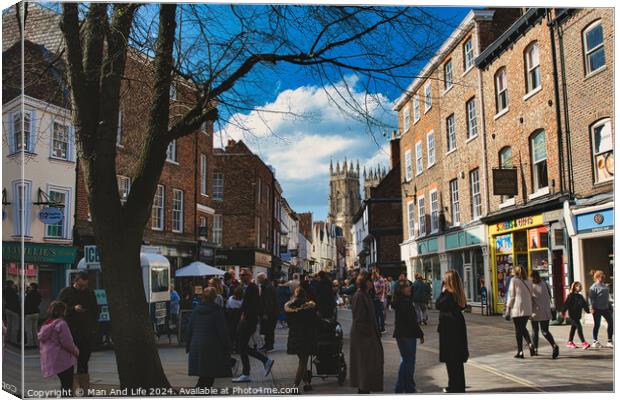 Bustling city street scene with pedestrians, historic buildings, and a cathedral spire under a blue sky with scattered clouds in York, North Yorkshire, England. Canvas Print by Man And Life