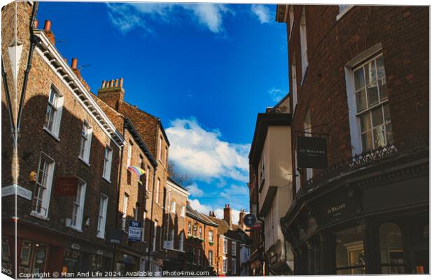 Charming European street scene with historic brick buildings under a clear blue sky with fluffy clouds, showcasing architectural details and local businesses in York, North Yorkshire, England. Canvas Print by Man And Life