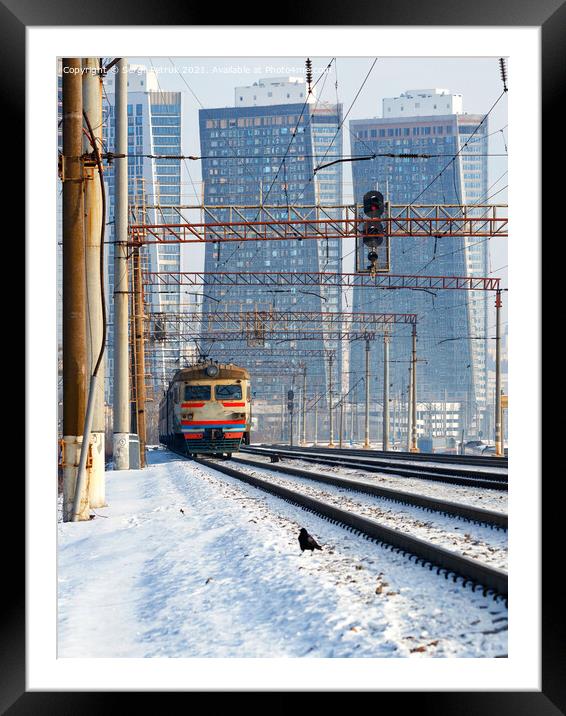 An electric train moves on rails against the backdrop of a cityscape of skyscrapers in a winter haze. Framed Mounted Print by Sergii Petruk