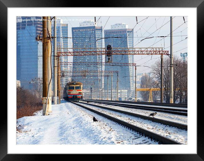 An old electric train moves on rails against the backdrop of a cityscape of skyscrapers on a sunny winter day. Framed Mounted Print by Sergii Petruk