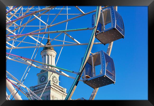 Empty cabins of the city's ferris wheel against the background of the blue sky and the spire of the old tower. Framed Print by Sergii Petruk