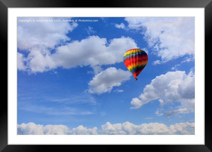 A motley multicolored hot air balloon raises a basket with tourists in the blue sky among white clouds. Framed Mounted Print by Sergii Petruk