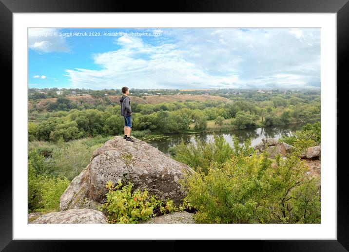 The teenager stands on top of a large stone boulder on the bank of the Southern Bug River and looks at the river below Framed Mounted Print by Sergii Petruk