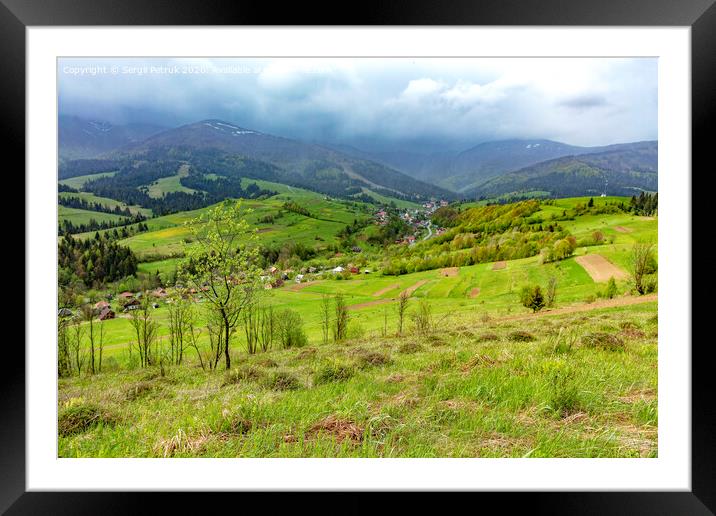 The mountain slope is covered with thick green grass against the backdrop of the picturesque landscape of the Carpathian Mountains shrouded in mist and villages in the valley. Framed Mounted Print by Sergii Petruk