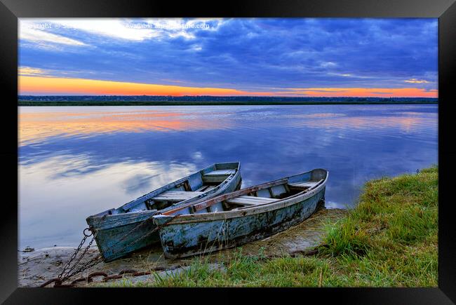Two old blue-green boats moored by a metal chain to the shore of a calm river against the backdrop of the bright rising sun. Framed Print by Sergii Petruk