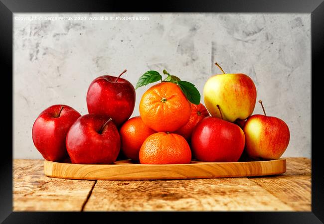 Red ripe apples and tangerines with green leaves lie on a wooden tray on an old wooden table with gray concrete background. Framed Print by Sergii Petruk