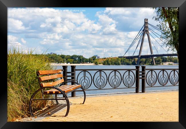 A wooden bench on the embankment of the Dnipro River against the background of yellow paving slabs and the northern bridge over the river in blur. Framed Print by Sergii Petruk