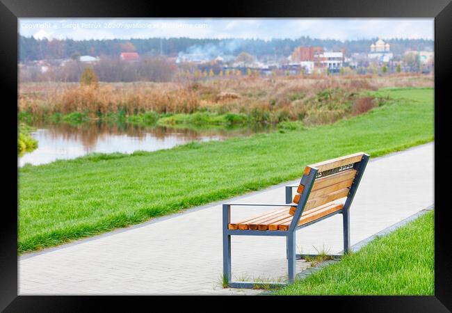 A wooden bench stands on a walking path along a beautiful green lawn on the river embankment with a blurred background of a rural landscape. Framed Print by Sergii Petruk