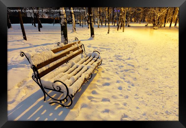 A wooden bench in a winter city evening park is covered with snow and illuminated by the warm light of a street lamp. Framed Print by Sergii Petruk